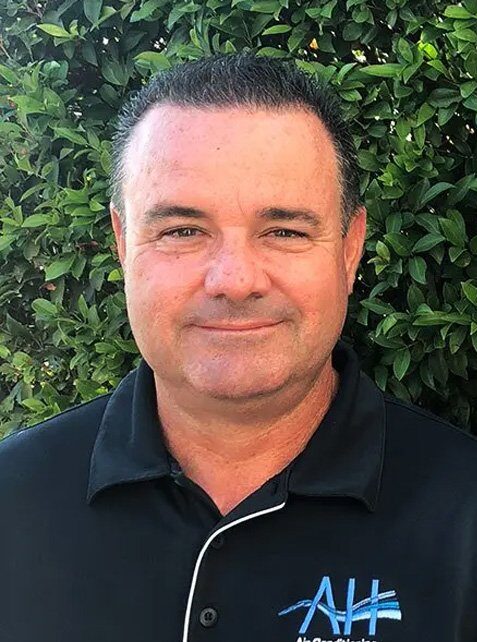 Portrait of a professional in a black AH Air Conditioning polo shirt, smiling in front of lush green foliage, representing the experienced team dedicated to customer care and tailored air conditioning solutions.